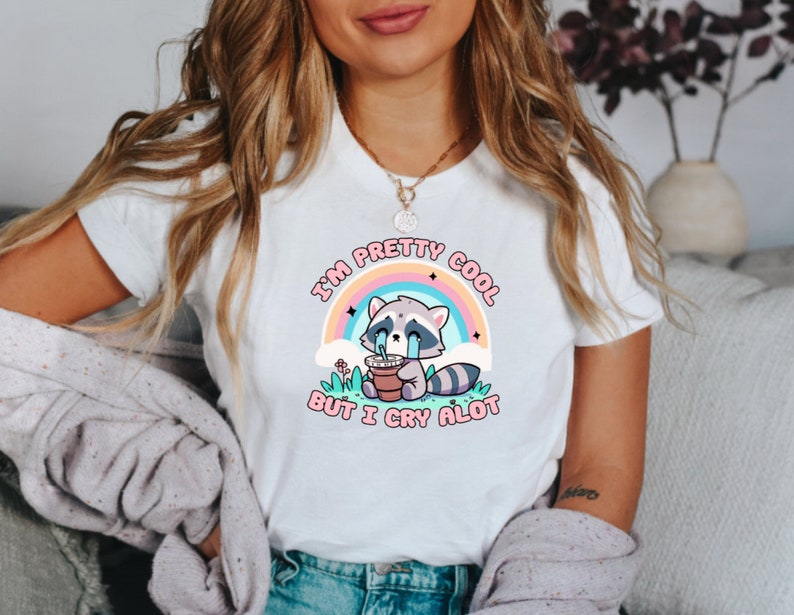 May include: White t-shirt with a graphic of a crying raccoon holding a cup of coffee. The text on the shirt reads "I'm pretty cool but I cry alot".