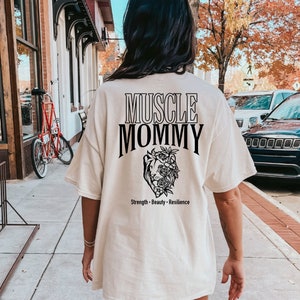 May include: A white t-shirt with black text that reads "Muscle Mommy" and a black floral design with the words "Strength - Beauty - Resilience" below.