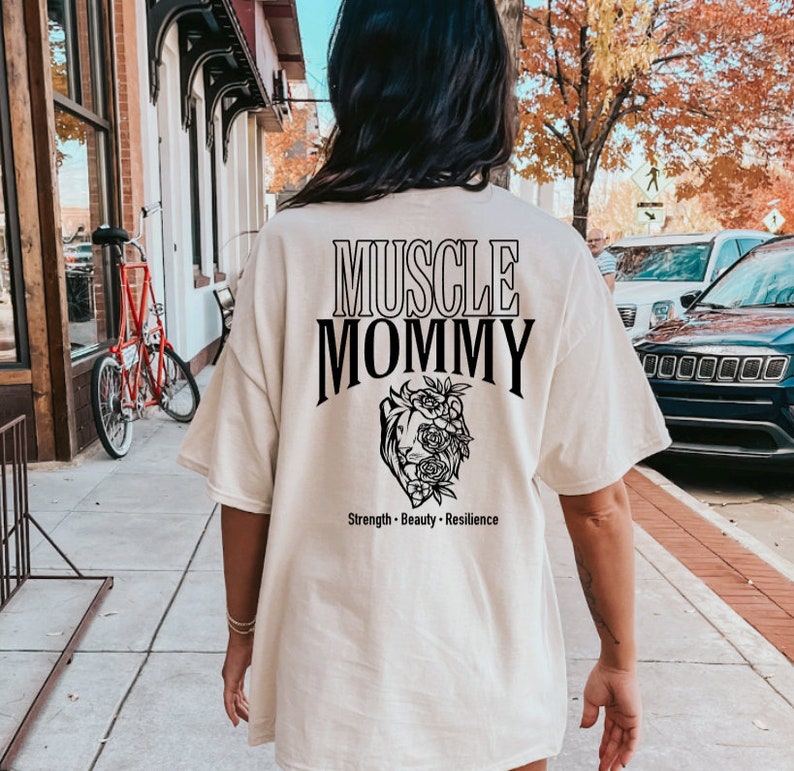 May include: A white t-shirt with black text that reads "Muscle Mommy" and a black floral design with the words "Strength - Beauty - Resilience" below.