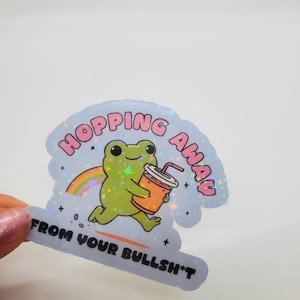 May include: A holographic sticker with a green frog holding a cup of orange juice. The sticker says "Hopping Away From Your Bullsh*t" in pink letters.