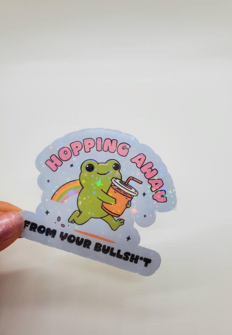 May include: A holographic sticker with a green frog holding a cup of orange juice. The sticker says "Hopping Away From Your Bullsh*t" in pink letters.