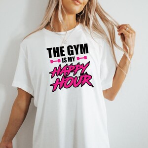 Peut inclure: T-shirt blanc avec un texte noir qui dit "The Gym is my Happy Hour" avec des ic&ocirc;nes de halt&egrave;res roses et du texte rose pour "Happy Hour".