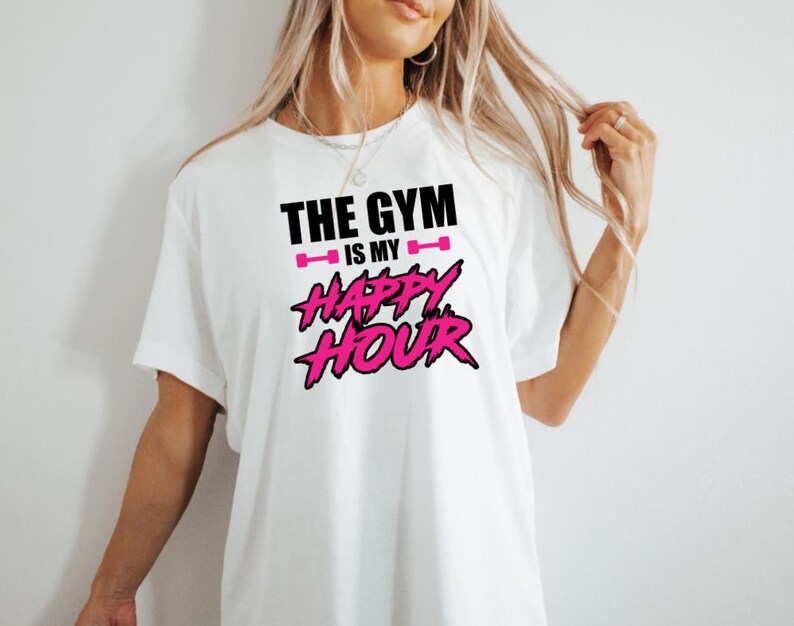Peut inclure: T-shirt blanc avec un texte noir qui dit "The Gym is my Happy Hour" avec des ic&ocirc;nes de halt&egrave;res roses et du texte rose pour "Happy Hour".