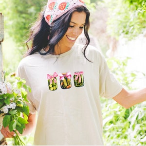 Pode incluir: Uma camiseta branca com um gr&aacute;fico de tr&ecirc;s potes de picles amarrados com fitas cor-de-rosa. Os potes est&atilde;o cheios de picles verdes e o fundo &eacute; uma imagem desfocada de folhagem verde.