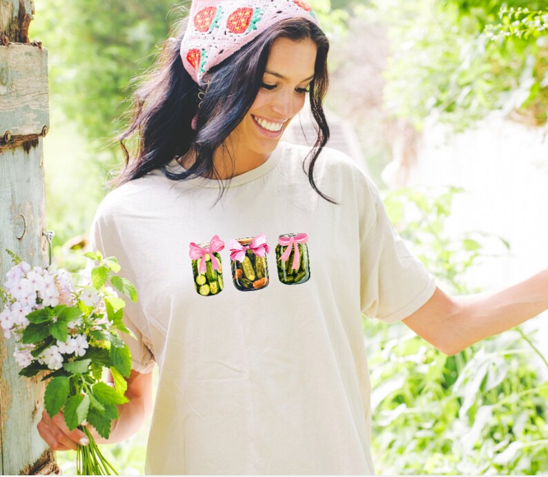 Pode incluir: Uma camiseta branca com um gr&aacute;fico de tr&ecirc;s potes de picles amarrados com fitas cor-de-rosa. Os potes est&atilde;o cheios de picles verdes e o fundo &eacute; uma imagem desfocada de folhagem verde.