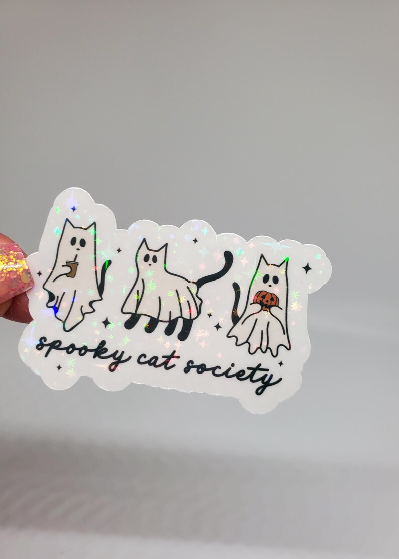 May include: A holographic sticker with three cartoon cats dressed as ghosts. The cats are holding a cup of coffee, a tail, and a pumpkin. The text "spooky cat society" is written below the cats.