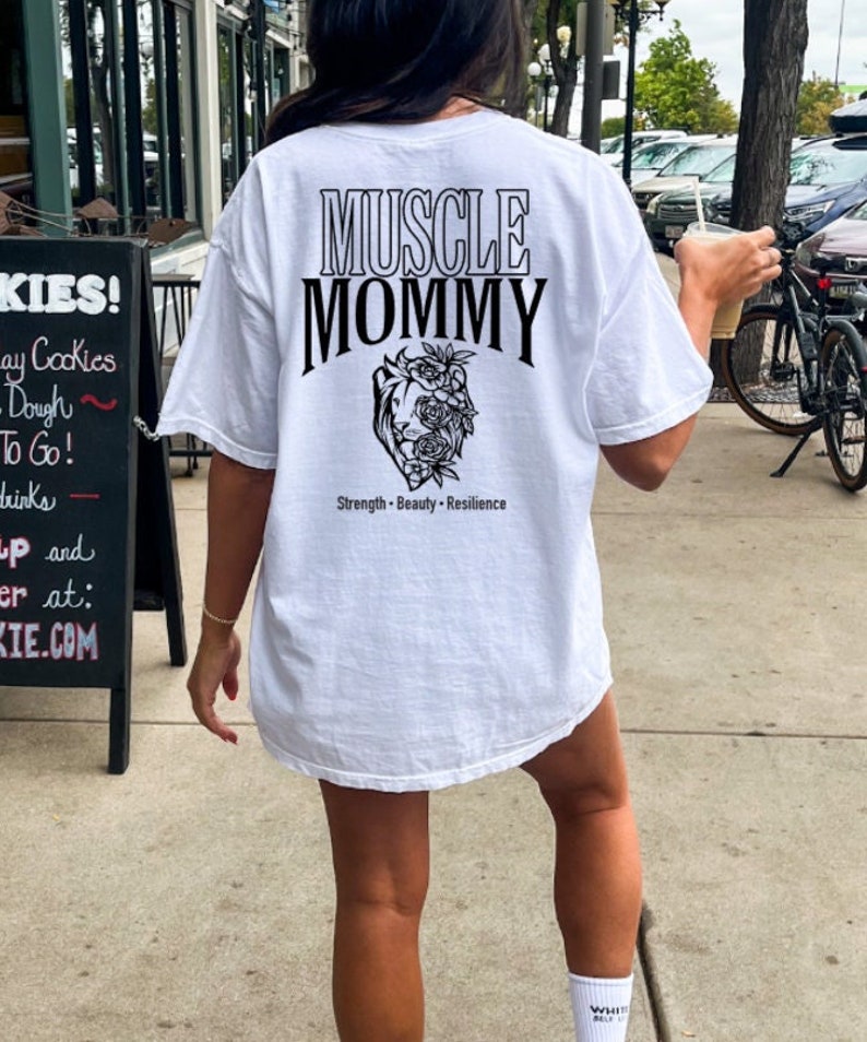 May include: White t-shirt with black text that reads "Muscle Mommy" and a floral graphic. The text below the graphic reads "Strength - Beauty - Resilience".