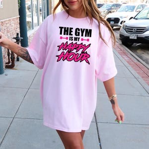 Peut inclure: T-shirt rose surdimensionn&eacute; avec le texte "The Gym is my Happy Hour" en lettres noires et roses.