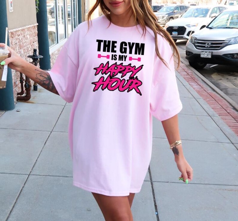 Peut inclure: T-shirt rose surdimensionn&eacute; avec le texte "The Gym is my Happy Hour" en lettres noires et roses.
