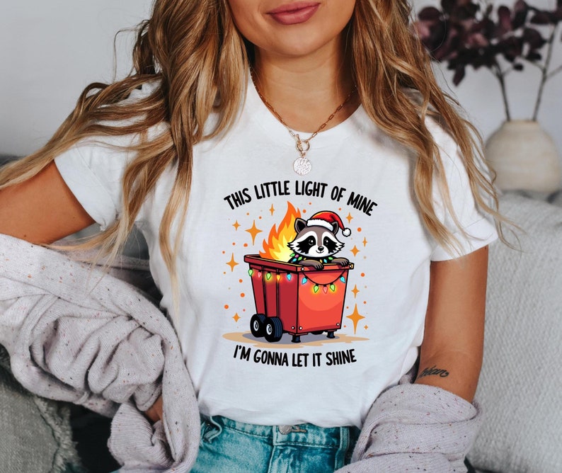 May include: White t-shirt with a graphic of a raccoon wearing a Santa hat in a burning dumpster with Christmas lights. The text on the shirt reads "This little light of mine I'm gonna let it shine."