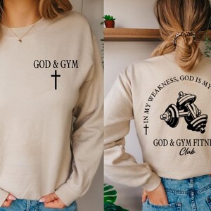 May include: A beige sweatshirt with the text "GOD & GYM" and a cross on the front. The back of the sweatshirt has the text "IN MY WEAKNESS, GOD IS MY STRENGTH" with a cross on each side and a dumbbell graphic in the center. The text "GOD & GYM FITNESS Club" is below the graphic.