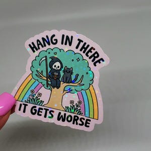 Op de afbeelding: Een roze holografische sticker met een cartoon afbeelding van de Grim Reaper die op een boom zit met een kat. De sticker heeft een regenboog en de tekst "Hang in there it gets worse".