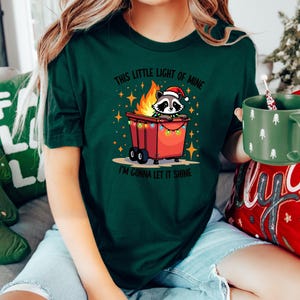 May include: A green t-shirt with a Christmas-themed design featuring a raccoon wearing a Santa hat in a burning trash can with Christmas lights. The text on the shirt reads "This little light of mine, I'm gonna let it shine."