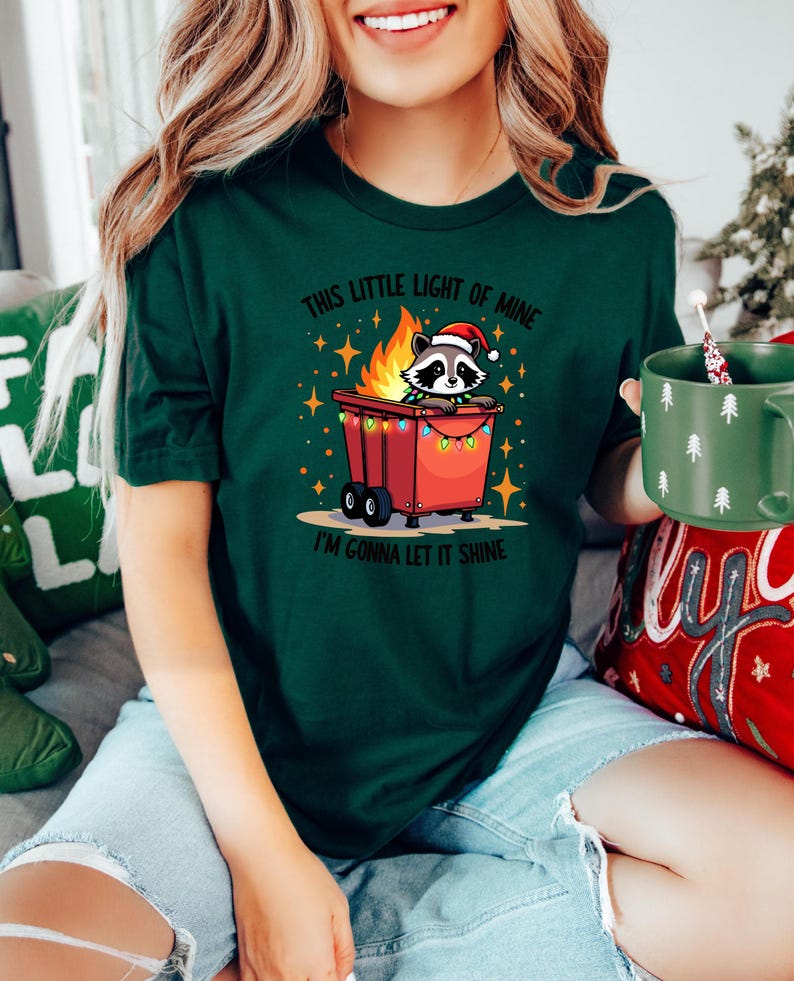May include: A green t-shirt with a Christmas-themed design featuring a raccoon wearing a Santa hat in a burning trash can with Christmas lights. The text on the shirt reads "This little light of mine, I'm gonna let it shine."