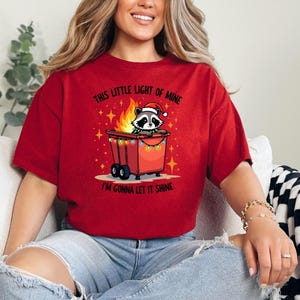 May include: Red t-shirt with a graphic of a raccoon in a trash can with Christmas lights. The text on the shirt reads "This little light of mine I'm gonna let it shine."