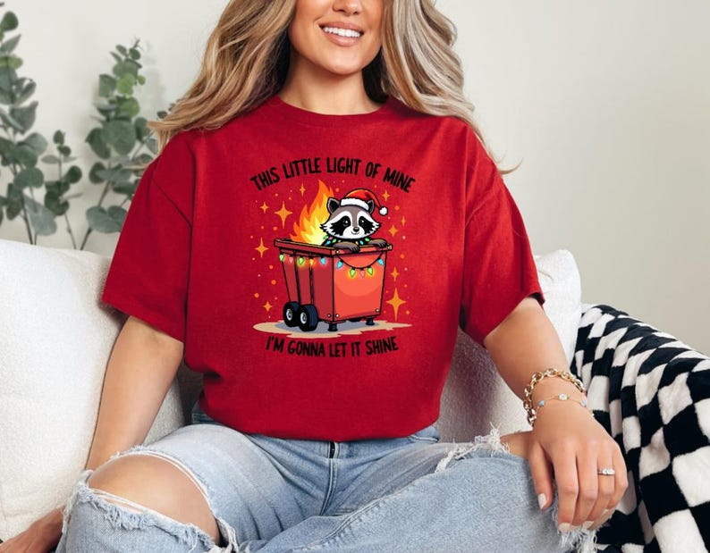 May include: Red t-shirt with a graphic of a raccoon in a trash can with Christmas lights. The text on the shirt reads "This little light of mine I'm gonna let it shine."