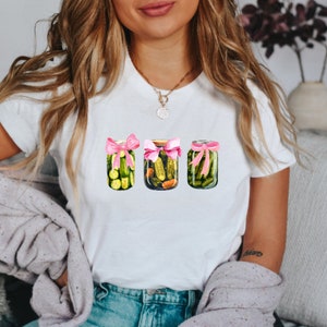 Pode incluir: Camiseta branca com um gr&aacute;fico de tr&ecirc;s frascos de picles amarrados com fitas cor-de-rosa.