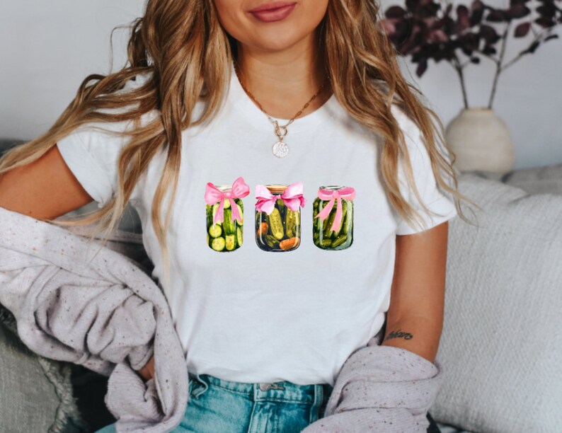 Pode incluir: Camiseta branca com um gr&aacute;fico de tr&ecirc;s frascos de picles amarrados com fitas cor-de-rosa.