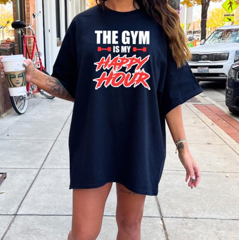 Peut inclure: Un t-shirt noir surdimensionn&eacute; avec le texte "The Gym is my Happy Hour" en lettres rouges et blanches. Le texte est stylis&eacute; avec un graphique de halt&egrave;re rouge et blanc de chaque c&ocirc;t&eacute; du texte "is my".