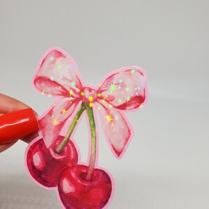 May include: A sticker depicting two red cherries with green stalks and a pink bow. The bow and cherries have a holographic effect, with sparkling stars. The sticker is set against a white backdrop.