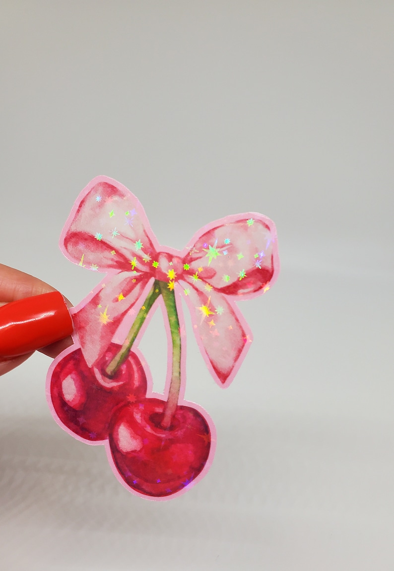 May include: A sticker depicting two red cherries with green stalks and a pink bow. The bow and cherries have a holographic effect, with sparkling stars. The sticker is set against a white backdrop.
