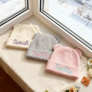 May include: Three ribbed knit beanies with bear ears are displayed on a windowsill. The beanies are cream, grey, and pink, each embroidered with a name in a contrasting colour: Sarah, Sophia, and Emily. The hats are a cosy winter accessory.