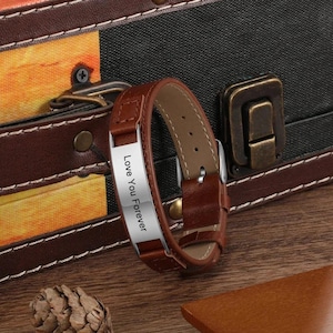 Puede incluir: Un brazalete de cuero marr&oacute;n con una placa plateada grabada con las palabras "Love You Forever".