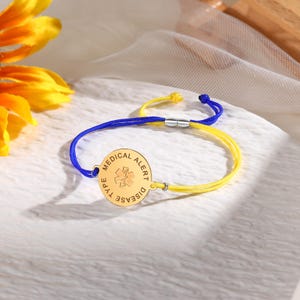 Puede incluir: Una pulsera de alerta médica de oro con un cordón azul y amarillo. La pulsera tiene una etiqueta redonda que dice "Tipo de enfermedad Alerta médica".