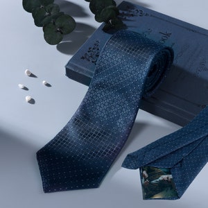 Puede incluir: Una corbata azul oscuro con estampado y un sutil efecto degradado. La corbata está enrollada y colocada encima de un libro azul con detalles dorados. El libro tiene la palabra "book" impresa en la portada.