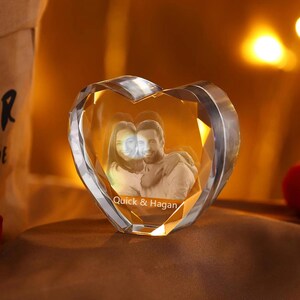 May include: A clear crystal heart-shaped paperweight with a 3D engraved image of a couple inside. The inscription "Quick & Hagan" is engraved at the bottom of the heart.