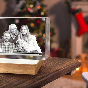 May include: A crystal cube with a family photo engraved inside. The cube is illuminated from below and sits on a wooden base. The photo shows a family of five, including two adults and three children. The photo is in black and white.