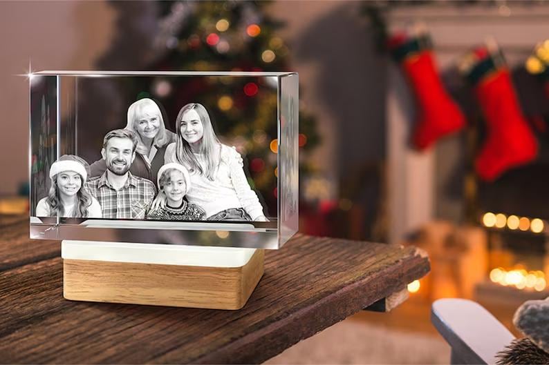 May include: A crystal cube with a family photo engraved inside. The cube is illuminated from below and sits on a wooden base. The photo shows a family of five, including two adults and three children. The photo is in black and white.