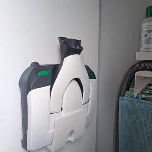 Puede incluir: Una aspiradora blanca y gris montada en una pared blanca. La aspiradora tiene un diseño curvo con un botón verde y un asa negra. La imagen está tomada desde un ángulo lateral, mostrando la forma plegada de la aspiradora.