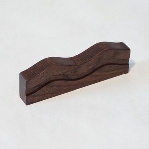 May include: A dark brown wooden stand with a wavy design. The stand has a groove in the middle, ideal for displaying small items.