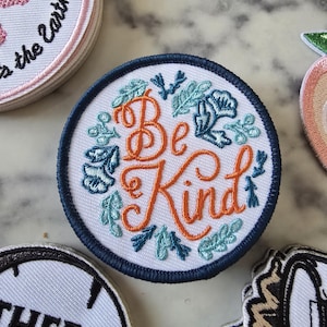 May include: A white embroidered patch with a navy blue border. The patch features a floral wreath with the words "Be Kind" in orange embroidery.