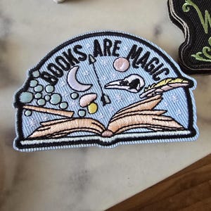 Puede incluir: Un parche bordado azul con el texto "Books Are Magic" en blanco. El parche presenta un libro abierto con una luna creciente, una calavera, una pluma y un hongo.