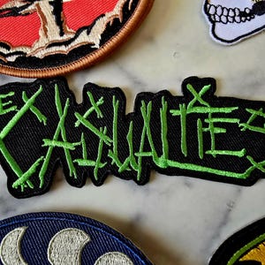 Può includere: Toppa ricamata nera con le parole "THE X-HALITES" in lettere verde neon stilizzate. La toppa è rettangolare con un bordo frastagliato. Altre toppe con diversi disegni sono visibili sullo sfondo.