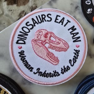 Puede incluir: Un parche redondo blanco bordado con puntadas rosas. El parche presenta una calavera de dinosaurio rosa con el texto "Dinosaurs Eat Man, Woman Inherits the Earth".