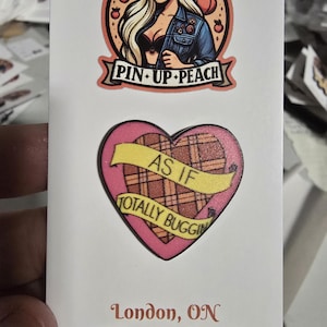 Op de afbeelding: Twee emaillepins. De eerste pin is een ronde, perzikkleurige pin met een cartoonvrouw met blond haar die een spijkerjack en een zwarte bh draagt. De tekst "Pin-Up Peach" staat onderaan de pin. De tweede pin is een roze hartvormige pin met een geel geruit patroon en een geel lint met de tekst "As If Totally Buggin".