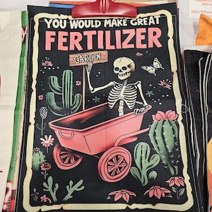 Puede incluir: Una funda de cojín negra con un diseño temático de jardín. El diseño presenta un esqueleto en una carretilla rosa con el texto "YOU WOULD MAKE GREAT FERTILIZER" y "GARDEN". El diseño incluye cactus, flores y una mariposa.