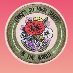 May include: Embroidered patch with a floral design. The patch features a central bouquet of red, white, blue, and purple flowers. The text "THERE'S SO MUCH BEAUTY IN THE WORLD" is embroidered around the edge in a circular pattern.