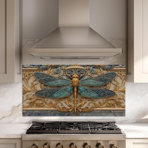 May include: A rectangular kitchen splashback panel featuring a large, detailed dragonfly with iridescent teal and blue wings centred against an ornate, raised gold-coloured relief pattern with scrolling filigree elements on a textured background.