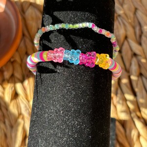 May include: Two colorful beaded bracelets. The top bracelet is clear with light green and blue beads. The bottom bracelet is multi-colored with gummy bear shaped beads in pink, blue, yellow, and green.