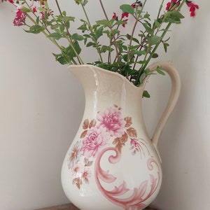 Op de afbeelding: Een witte keramische kan met een bloemenpatroon in roze en bruine tinten. De kan heeft een brede opening en een handvat. Hij is gevuld met een boeket roze en rode bloemen.
