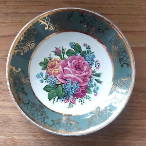 May include: Vintage decorative plate with a teal border and gold filigree accents. The center features a floral bouquet of pink and yellow roses, with blue forget-me-nots. The plate has a gold rim and is likely made of porcelain.