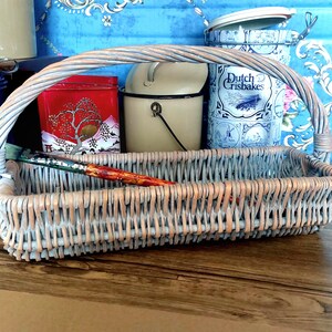 May include: A gray wicker basket with a handle, sitting on a wooden surface. The basket contains a paintbrush, a red tin with a Japanese scene, a white enamelware container, and a blue tin with the text "Dutch Crispbakes".