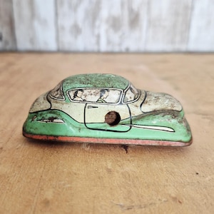 May include: Vintage toy car in green, white, and orange. The metal car features a detailed design with a depiction of two people inside. The car has a distressed, aged appearance, suggesting it is an antique collectible.