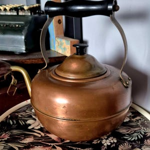 May include: A vintage copper kettle with a black wooden handle and a black knob on the lid. The kettle is sitting on a tray with a floral pattern.