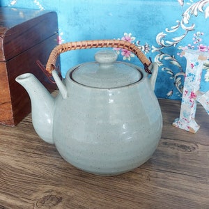 Può includere: Una teiera in ceramica grigio chiaro con un manico intrecciato marrone. La teiera ha una finitura macchiettata e un corpo arrotondato.