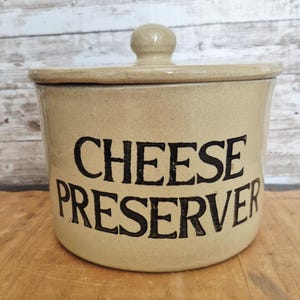 May include: Tan ceramic cheese preserver with a lid and a knob handle. The words "CHEESE PRESERVER" are printed in black, bold letters on the front of the container. The container is cylindrical and sits on a wooden surface.